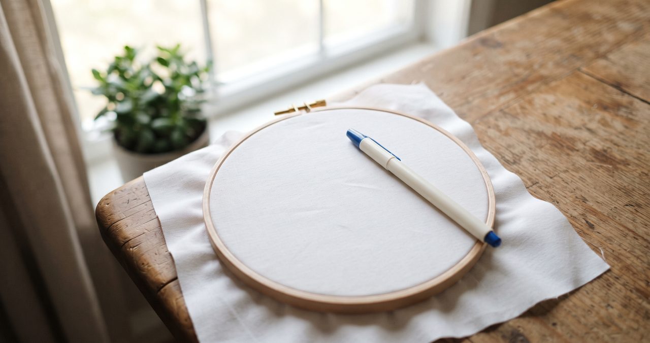 窓際のテーブルに丸い刺繍枠に張られた白い生地と布用チャコペンを置いた俯瞰の物撮り写真、人物なし