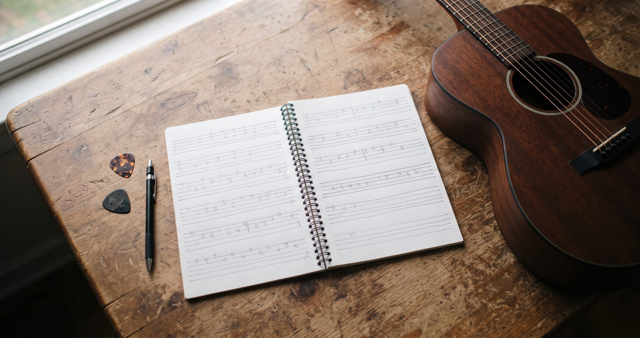 机の上に開いたノートに手描きの数字だけのタブ譜が書かれ、傍らにギターが置かれた俯瞰の静物、人物なし