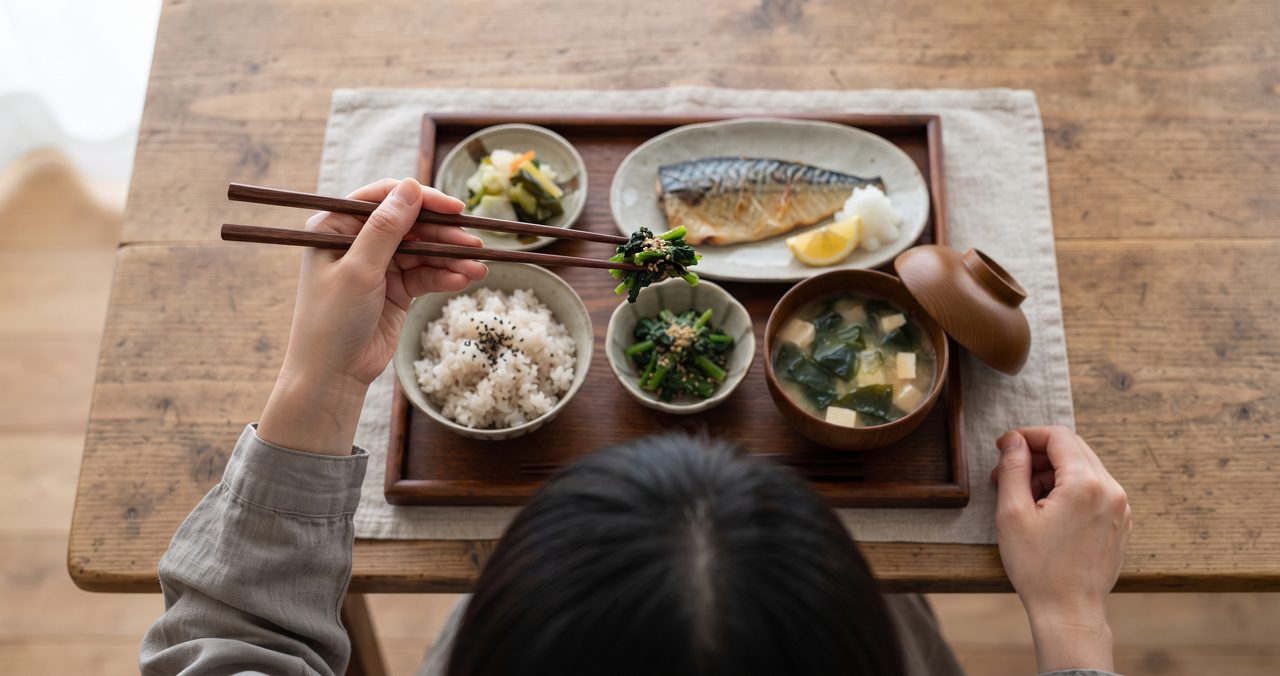 一人前の和食の食卓で左手に箸を持ち小鉢をつまむ俯瞰ショット、上の箸を親指・人差し指・中指でつまみ下の箸との間に中指が入り2本が明確に分離している、頭部が画面下端に写る構図